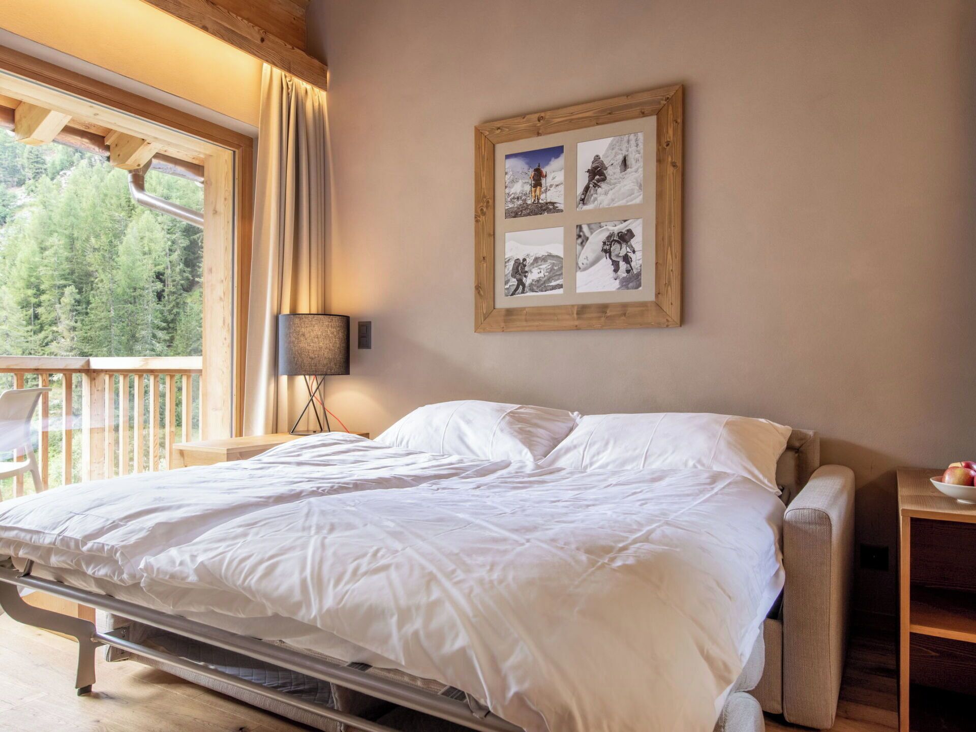 Un dormitorio con cama, mesita de noche, lámpara, collage de fotos enmarco y vista a árboles a través de una ventana.