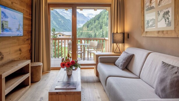 Ein gemütliches Zimmer mit Holzvertäfelung, einer Couch, einem Couchtisch mit Blumen, einem Fernseher an der Wand und einem Balkon mit Bergblick.