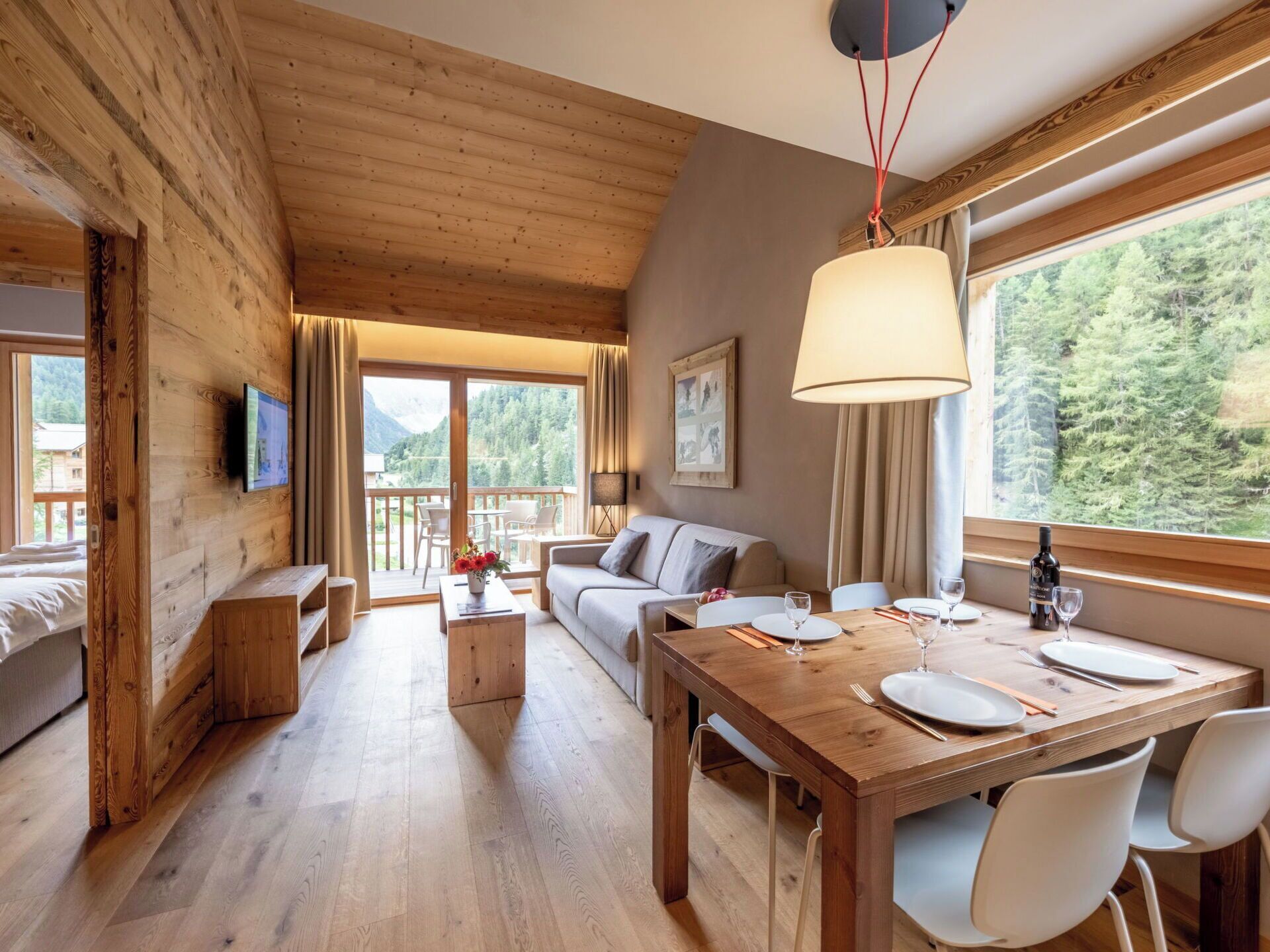 A wooden interior with a dining table set for four, a sofa, a TV mounted on the wall, and a window with a forest view.