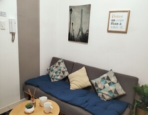 Ein Zimmer mit einer Couch, einem kleinen Tisch mit Tassen und einer Pflanze, einem gerahmten Bild des Eiffelturms und einem an der Wand montierten Telefon.