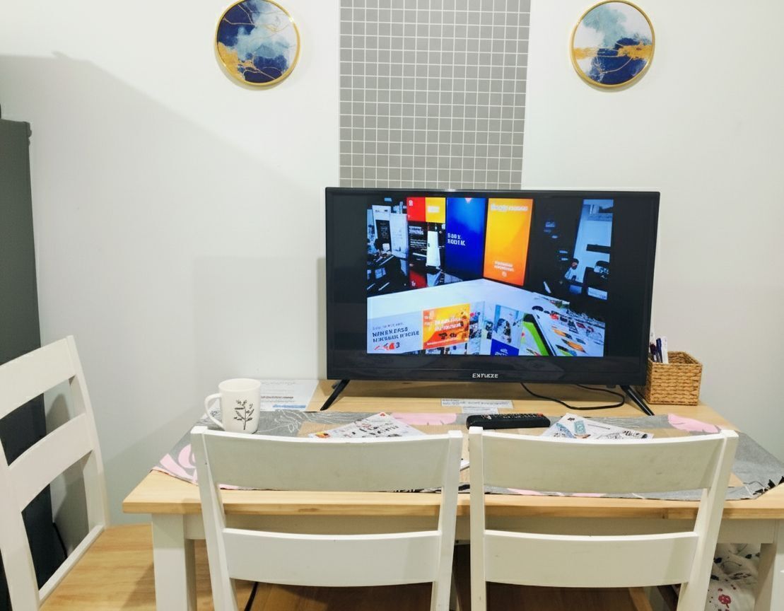 A television on a wooden table with two white chairs, a white mug, and a basket with pens.