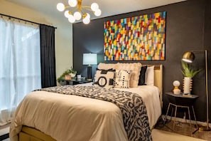 Un dormitorio moderno con cama, mesita de noche, lámpara y un cuadro abstracto de colores en la pared.