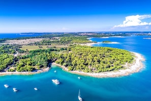 An aerial view of a coastal area with a small island surrounded by clear blue waters, several boats, and a distant cityscape. - Sun And Fun (Pula)