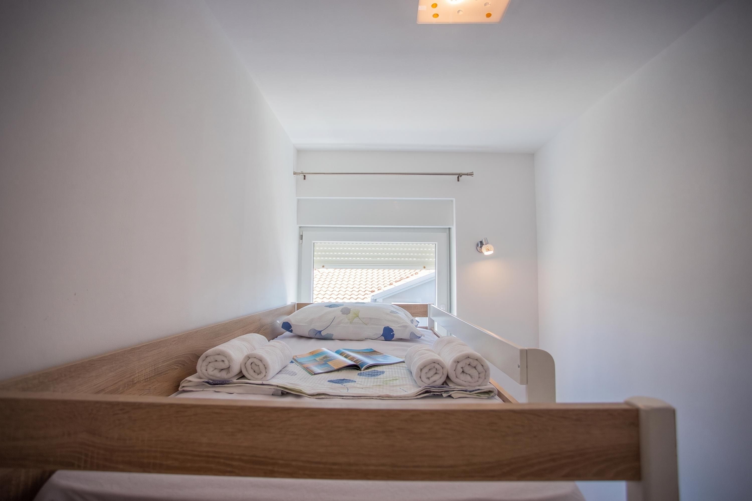 A small, neatly made bed with rolled towels and a book, in a room with white walls and a sloped ceiling.