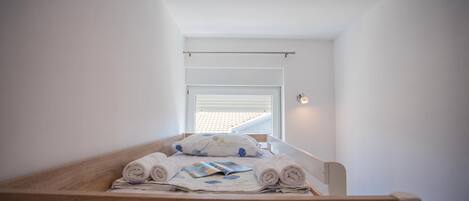 A small, neatly made bed with rolled towels and a book, in a room with white walls and a sloped ceiling.