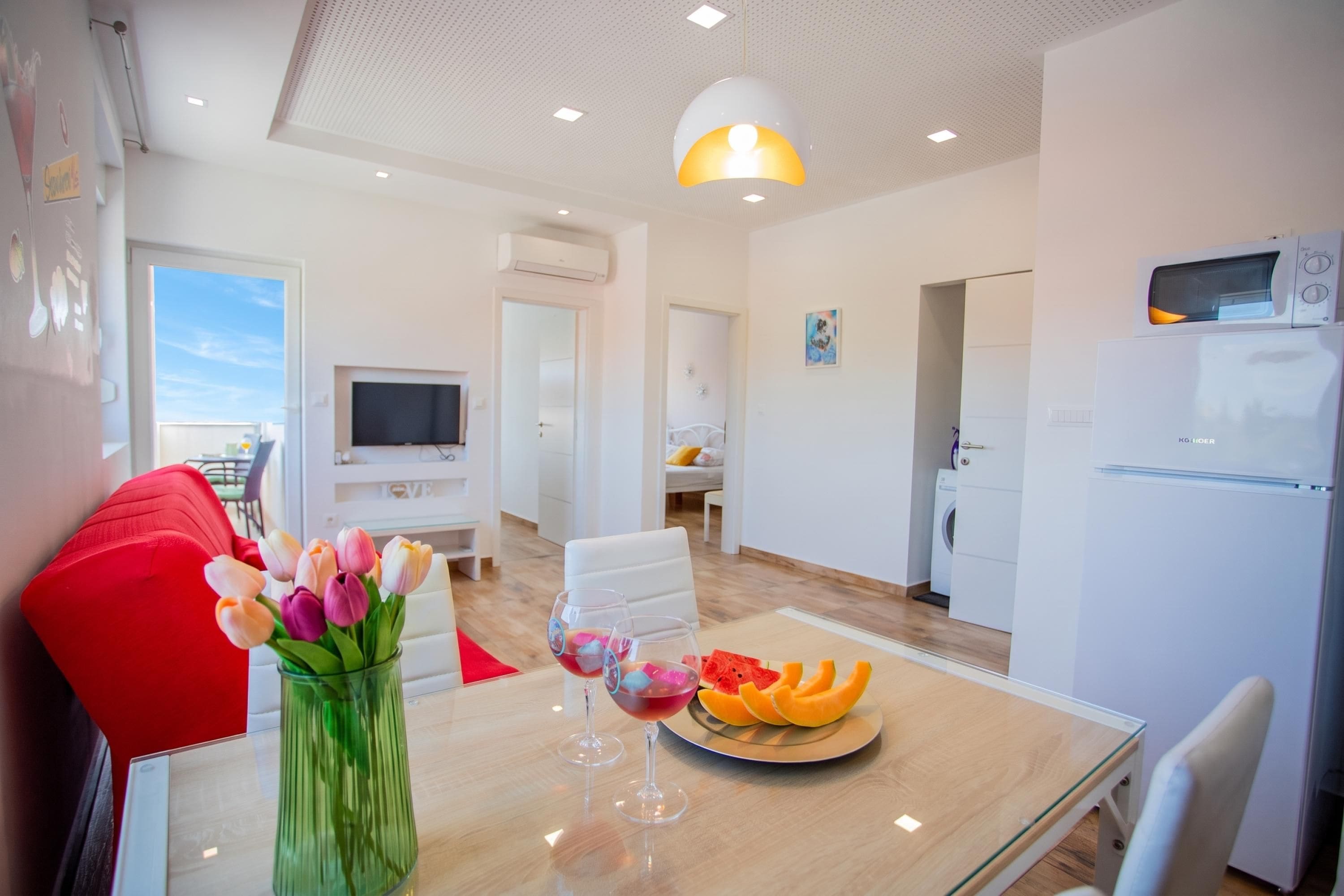 Una sala de estar moderna con un sofá rojo, una mesa de centro de vidrio y una disposición de frutas, además de un televisor montado en la pared.