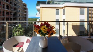 A balcony with a table, a vase with flowers, and a view of buildings and greenery. - 203 B, One Bedroom Apartment (Vlorë)