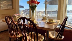A dining area with a table set for four, chairs, a vase with flowers, and a view of the sea through large windows. - Lakeside Paradise - Hot Tub, Dock, 5bdrm, Firepit (Victorville)