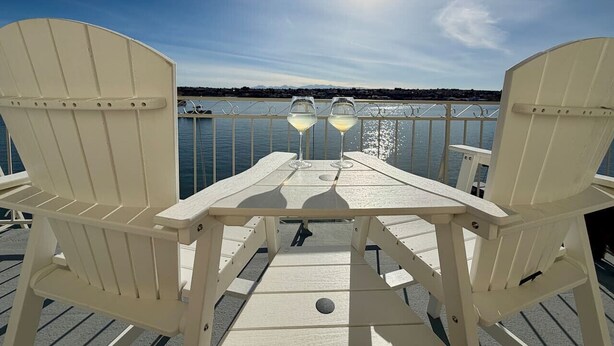 A deck with two white Adirondack chairs and a table, two glasses of white wine, and a view of the water and a boat in the background. - Lakeside Paradise - Hot Tub, Dock, 5bdrm, Firepit (Victorville)