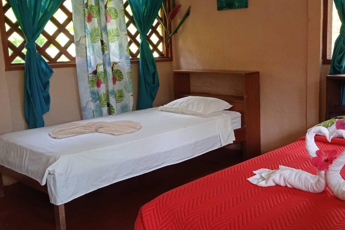 A room with two beds, one with a white bedspread and the other with a red bedspread. There are curtains with a leaf pattern, and a wooden headboard is visible.