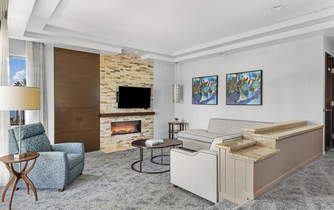 A modern hotel room with a stone fireplace, a flat-screen TV, a sofa, a coffee table, and artwork on the walls.