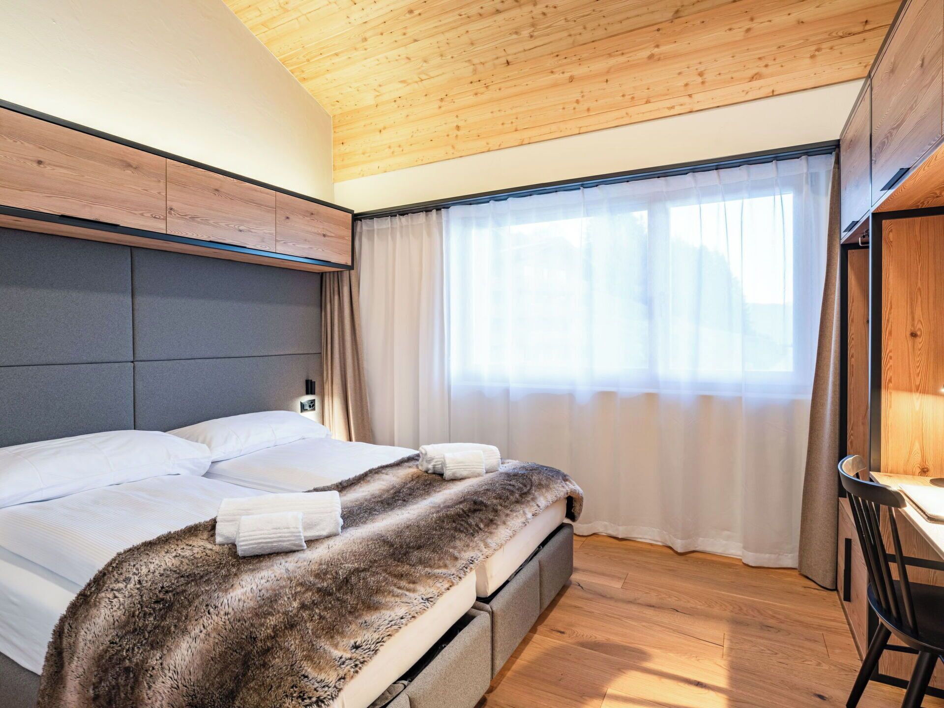 A modern bedroom with a wooden ceiling, a bed with a fur throw, a bedside table, and a window with curtains.