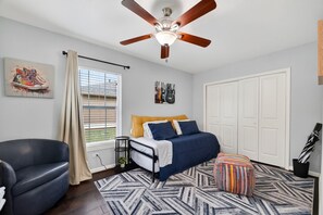 Ein Schlafzimmer mit Bett, Deckenventilator, Fenster mit Jalousien, Stuhl und Tür.