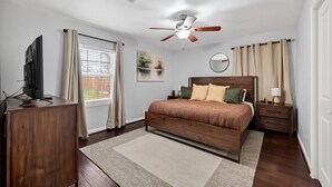 Ein Schlafzimmer mit Bett, Deckenventilator, Fernseher, Schreibtisch und einem Fenster mit Vorhängen.