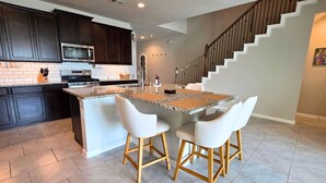 A modern kitchen with dark cabinets, a granite countertop, and a central island with bar stools. - Corporate Retreat Near Houston and Med Ctr (Pearland)