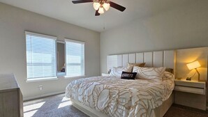 A bedroom with a large bed, a ceiling fan, a nightstand with a lamp, and a window with blinds. - Corporate Retreat Near Houston and Med Ctr (Pearland)
