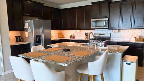 A kitchen with dark cabinets, a stainless steel refrigerator, a microwave, a sink, and a small trash bin. - Corporate Retreat Near Houston and Med Ctr (Pearland)
