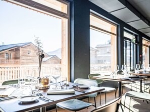 Ein Restaurant mit gedeckten Tischen, großen Fenstern mit Blick auf Gebäude und Berge sowie einem modernen Innenraum.