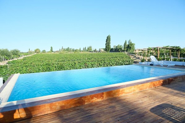 A rectangular pool with a wooden deck, surrounded by lush greenery and a clear sky.