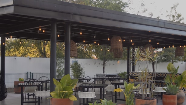 Un patio al aire libre con mesas y sillas, plantas en macetas y luces colgantes.