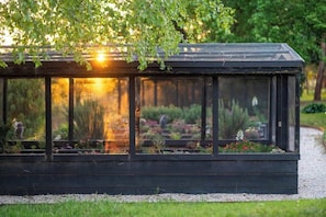 A glass-enclosed structure with a metal roof, surrounded by a garden with various plants and flowers. - 'Meadow Mist' Gardens plus Spa and Fire Pit (Far Meadow)