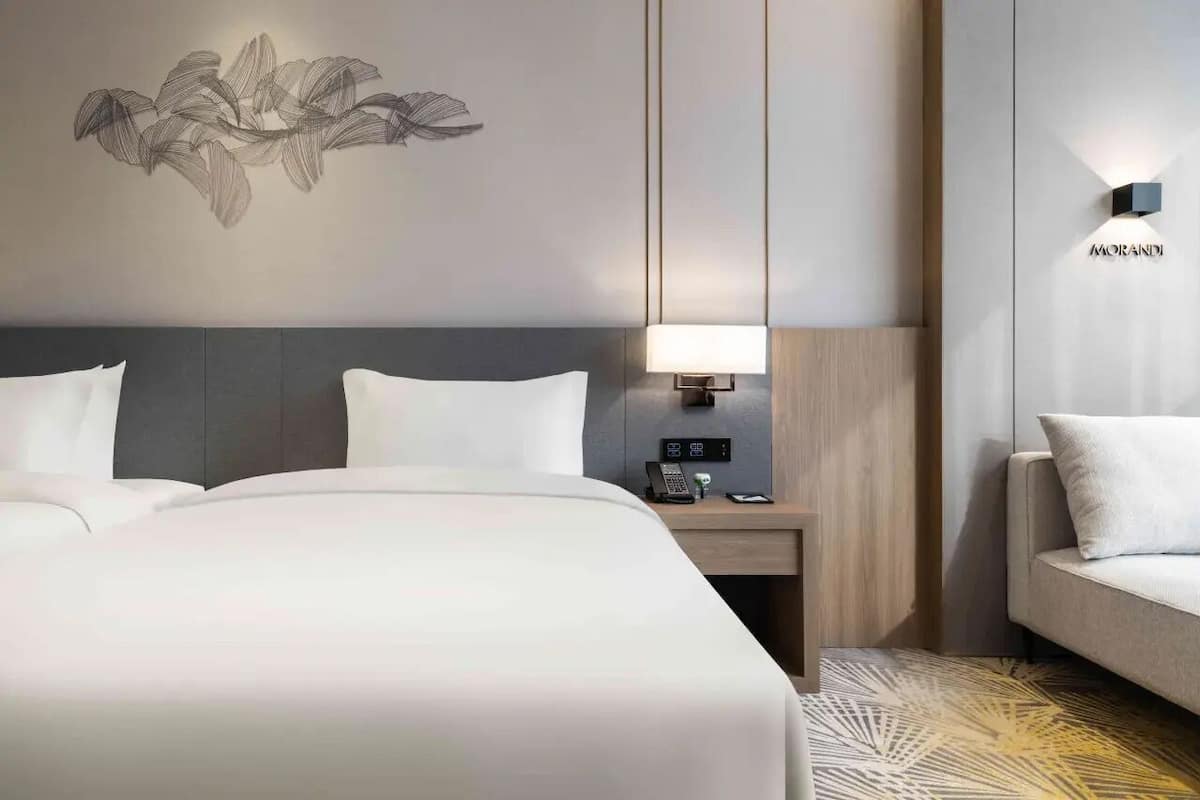 A hotel room with a large bed, bedside tables, a wall-mounted lamp, a sofa, and a decorative wall piece.