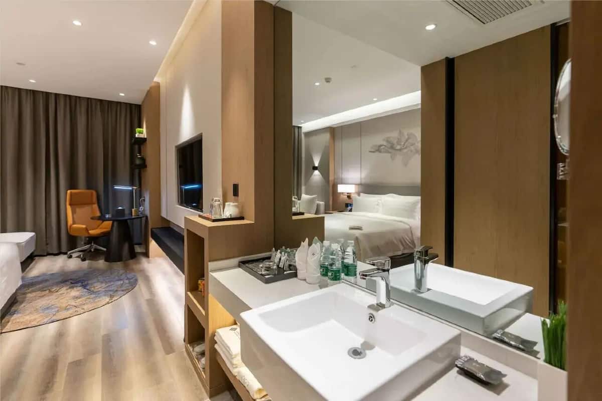 A modern hotel room with a large bed, a desk, a chair, a TV, a large mirror, and a bathroom with a double sink.