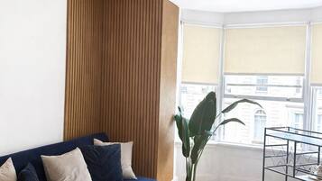 A living room with a blue sofa, a glass coffee table, a wooden accent wall, a potted plant, and a window with blinds.