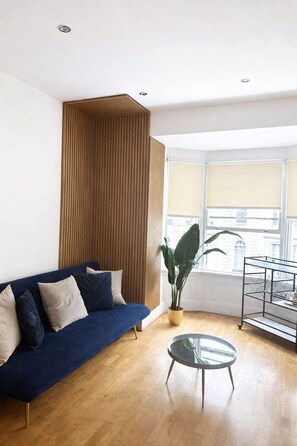 Ein Wohnzimmer mit blauem Sofa, einem Glastisch, einer Holz-Akzentwand, einer Topfpflanze und einem Fenster mit Jalousien.