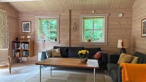 A cozy cabin interior with a wooden floor, a sofa, a coffee table, a bookshelf, and two windows with a view of greenery. - Traditional log house near beach and forest (Rørvig)