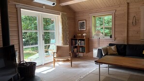A cozy cabin interior with a sofa, a wicker chair, a bookshelf, and a large window. - Traditional log house near beach and forest (Rørvig)