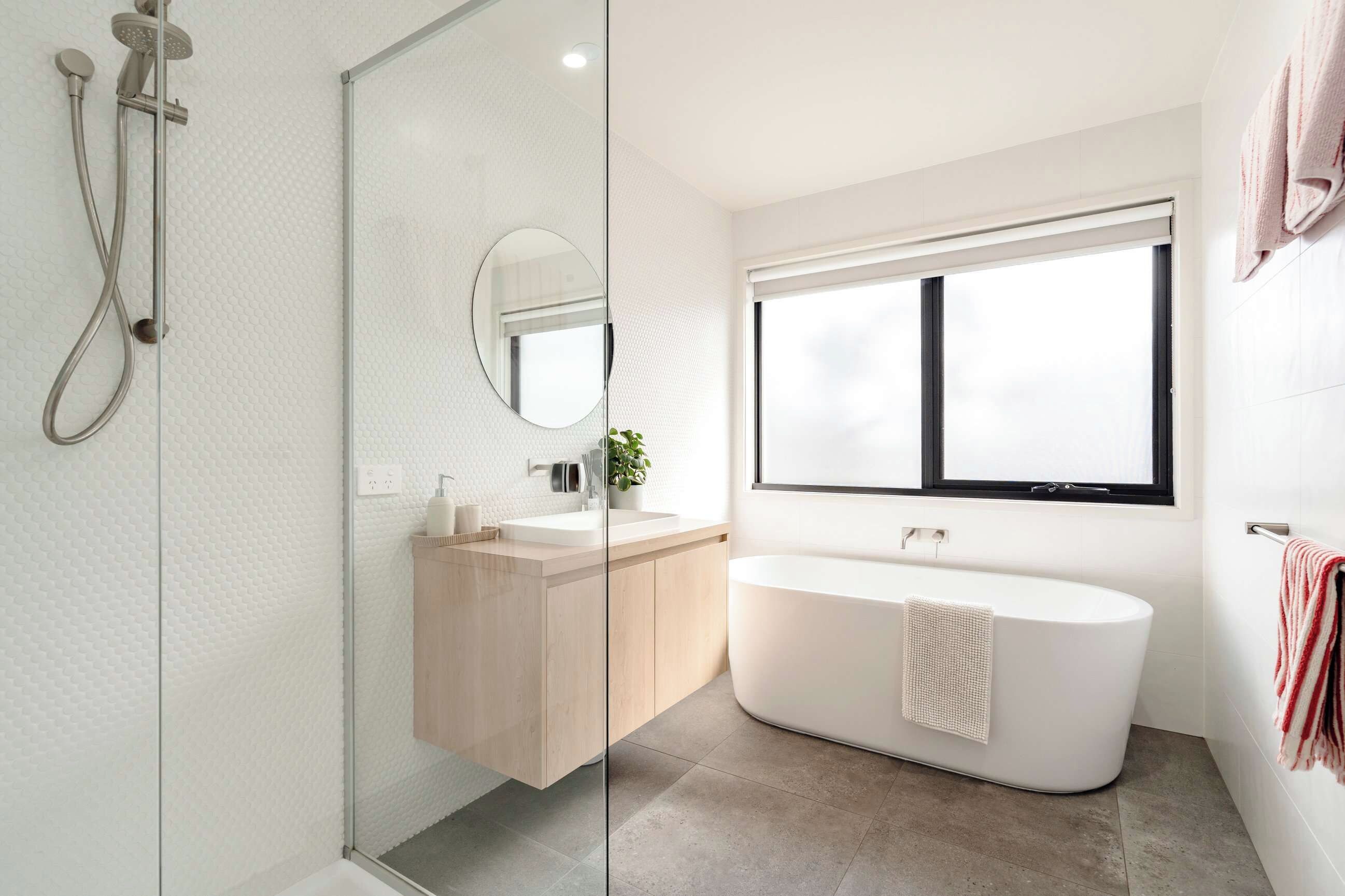 Une salle de bain moderne dotée d’une douche à parois de verre, d’une baignoire sur pied, d’un miroir rond et d’une fenêtre laissant entrer la lumière naturelle.