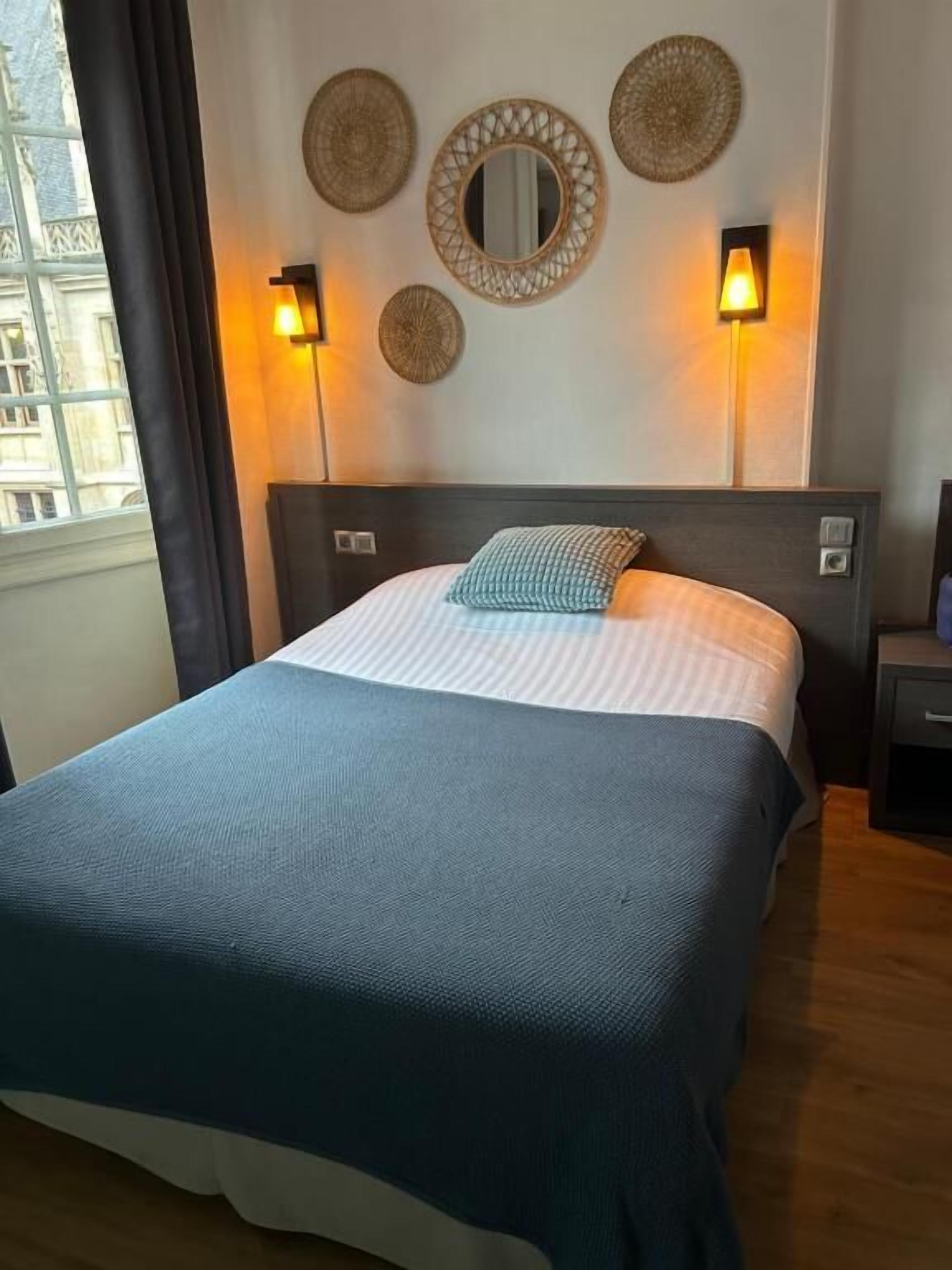 A hotel room with a bed, a headboard, two wall sconces, and decorative wall art.