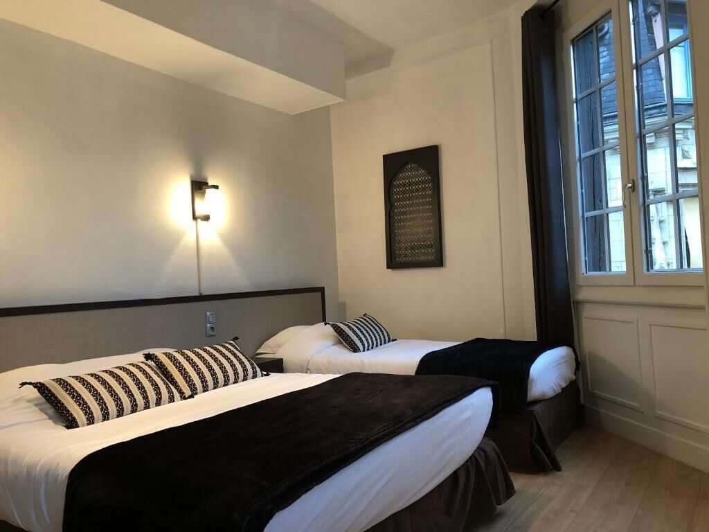 Quarto de hotel com duas camas, uma luminária fixada na parede, uma janela com persianas e um objeto decorativo na parede.