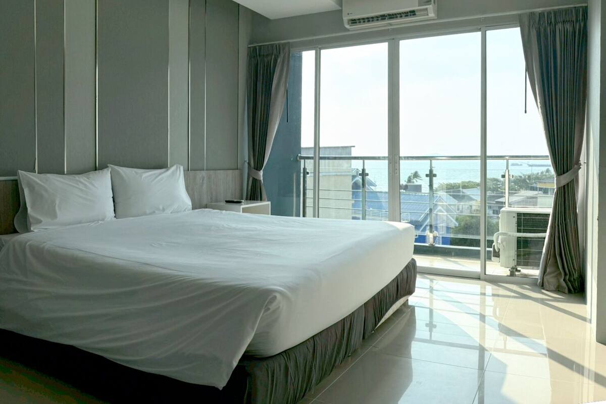 A hotel room with a large bed, a nightstand, an air conditioning unit, and a balcony with a view of buildings.