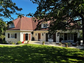 Une maison à deux étages avec un toit recouvert de tuiles rouges, une véranda à l’avant avec des sièges d’extérieur et une pelouse bien entretenue.