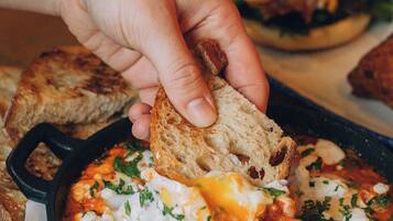 A hand dipping a piece of bread into a skillet with a dish that includes eggs and herbs.