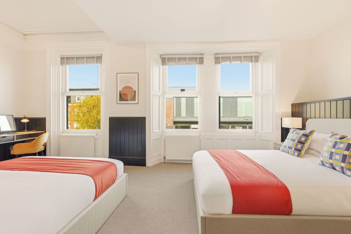 A hotel room with two beds, a desk, a chair, and a window with a view of buildings and trees.
