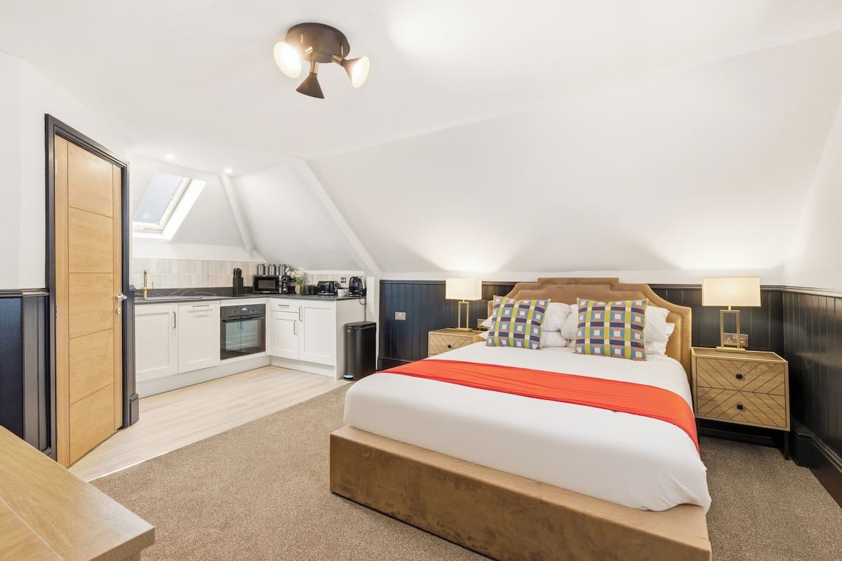 A bedroom with a bed, bedside tables, a nightstand, a window, a skylight, a kitchen area, and a door.