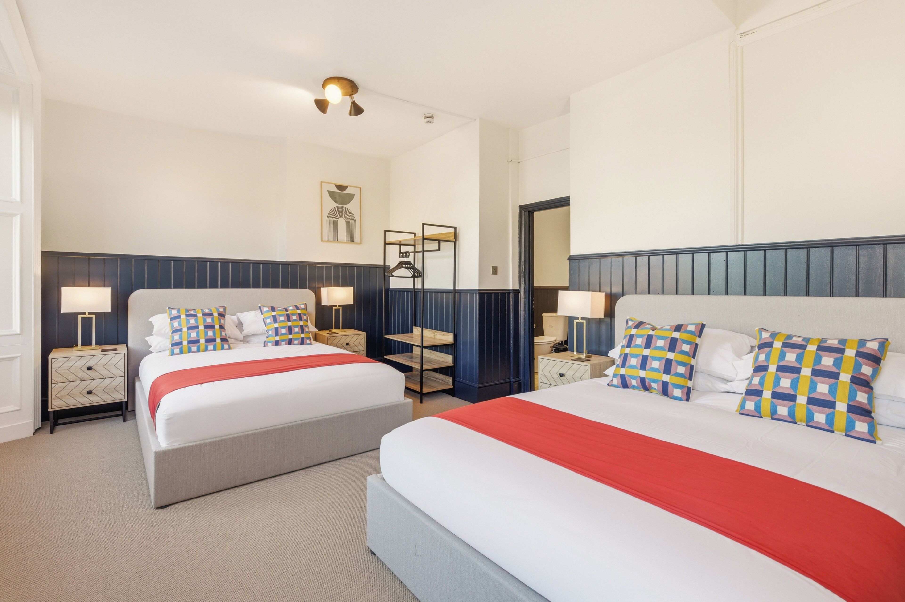Ein Hotelzimmer mit zwei Betten, jeweils mit roter Decke, weißer Bettwäsche und gemusterten Kissen. Es gibt Nachttische mit Lampen, ein Wandregal und einen Spiegel.