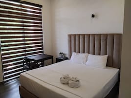 A hotel room with a bed, a bedside table, a small table, and a window with blinds.