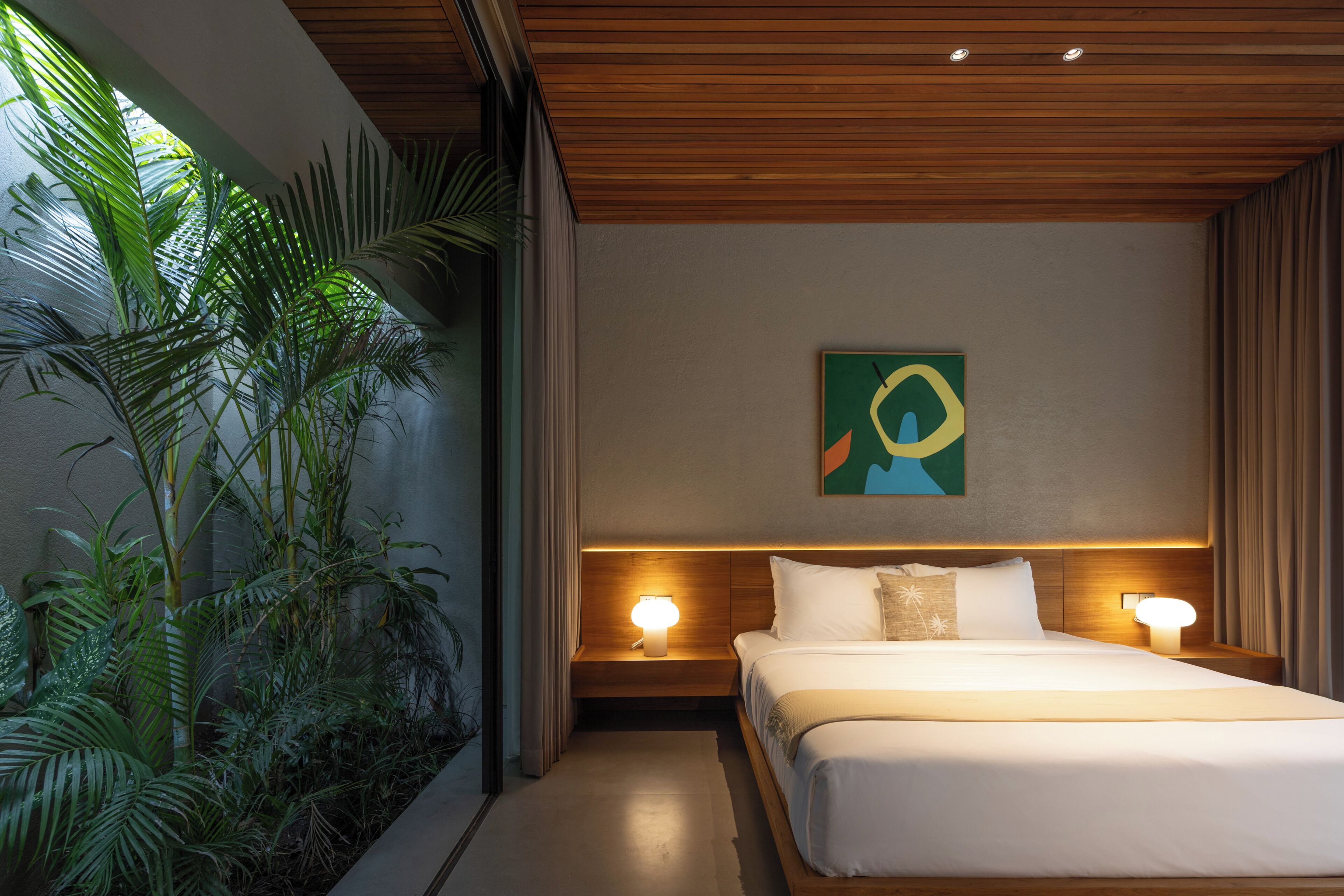 A hotel room with a bed, two bedside lamps, a wall-mounted artwork, and a large window with plants.