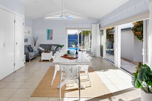 A spacious living room with a dining area, a ceiling fan, a large window, and a sliding glass door leading to a patio. - Endless Summer Sawtell - Luxe Beach Escape + Pool (Sawtell)