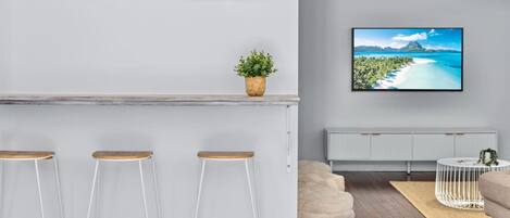 Un salon moderne doté d’un coin bar, de tabourets de bar blancs, d’un grand téléviseur à écran plat, d’une plante en pot et d’un espace salon confortable.