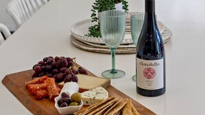 Une table à manger avec une bouteille de vin, une planche de fromages et une assiette de raisins.