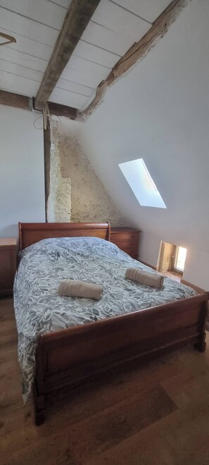 Ein Schlafzimmer mit schrägen Wänden, einem hölzernen Bett, einem Nachttisch und einem kleinen Fenster.