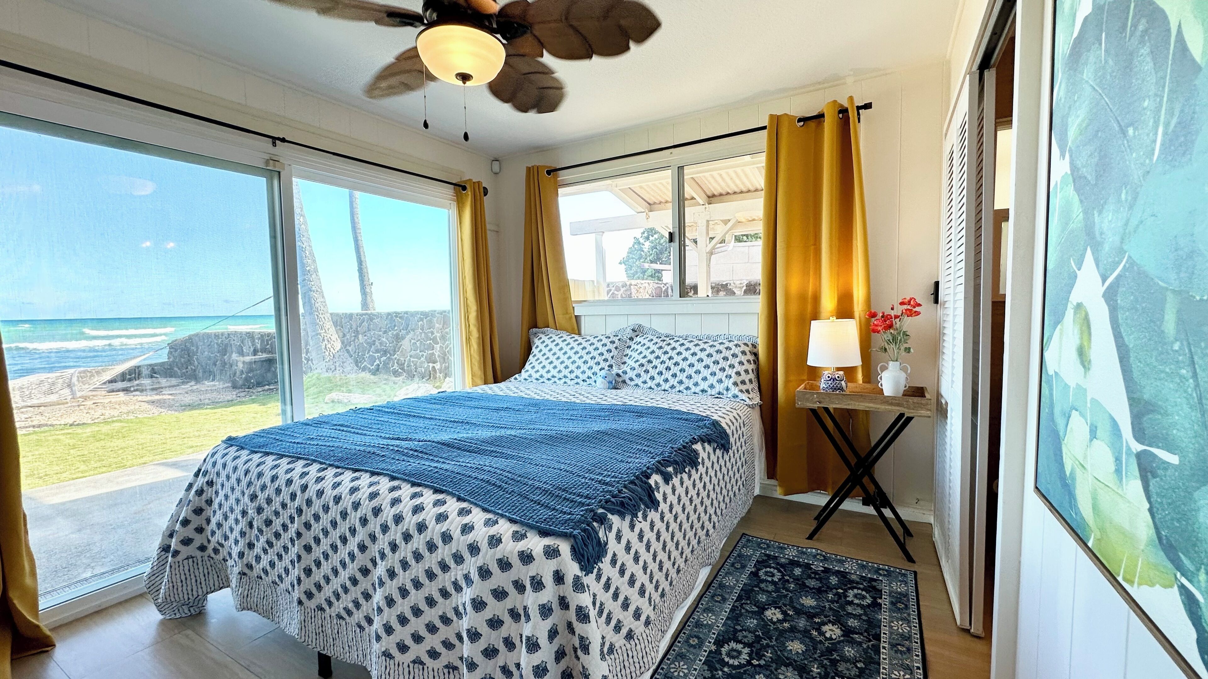 A bedroom with a bed, a nightstand, a lamp, a window with curtains, and a view of the ocean.