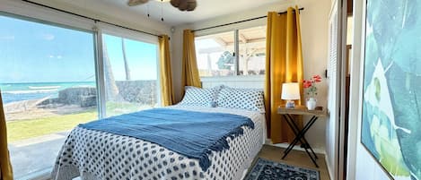 A bedroom with a bed, a nightstand, a lamp, a window with curtains, and a view of the ocean.