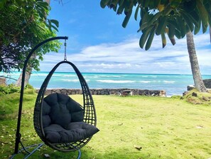 A hanging chair by a coastal view with a stone wall and palm trees. - Oceanfront 2BR/2BA Home · 2 Kings, Fast WiFi, AC, BBQ & Fire Pit (Ewa Beach)