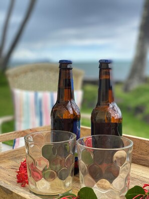 Two bottles on a wooden tray with glasses, surrounded by red flowers, with a blurred outdoor view in the background. - Oceanfront 2BR/2BA Home · 2 Kings, Fast WiFi, AC, BBQ & Fire Pit (Ewa Beach)
